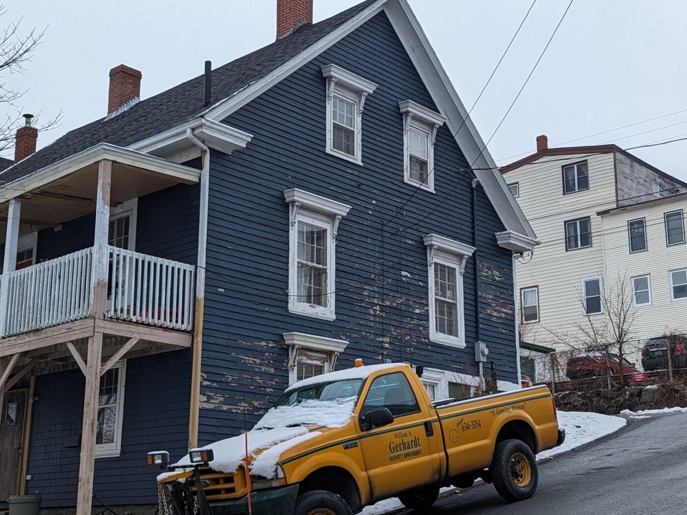 Yellow pickup truck with a snow plow parked in front of a navy blue wood-sided house in winter