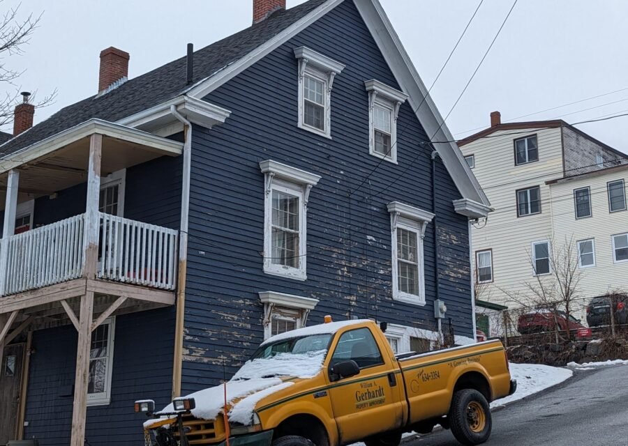 Yellow pickup truck with a snow plow parked in front of a navy blue wood-sided house in winter