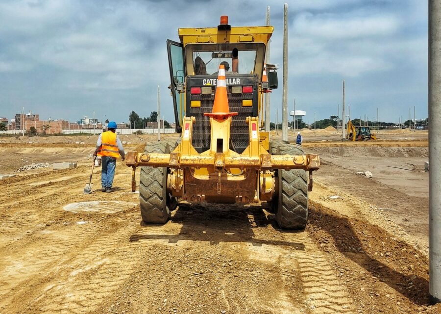 Yellow motor grader on a dirt construction site with a worker in a safety vest walking nearby